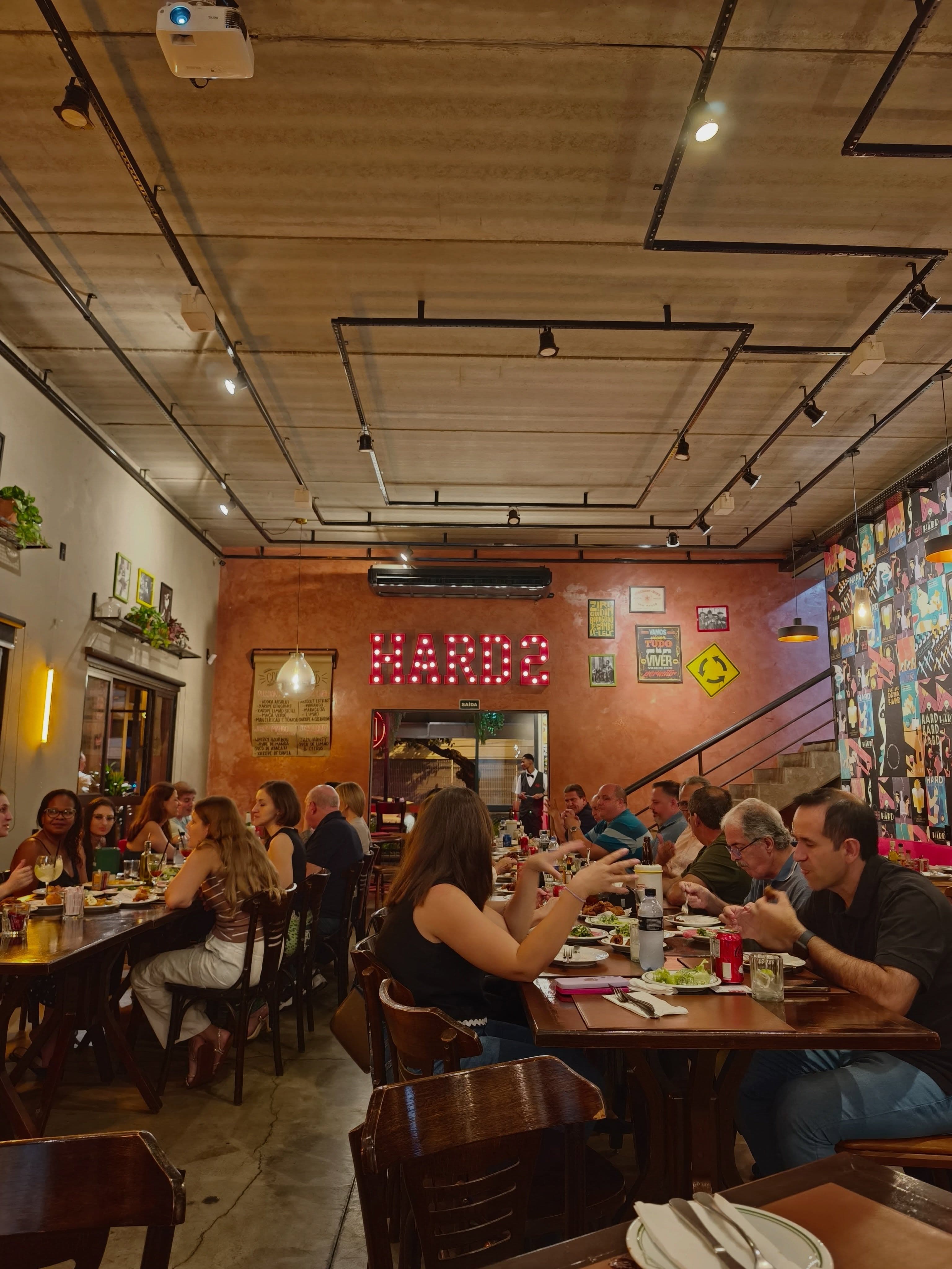 Salão do Hard Chopp 2 cheio, com o letreiro HARD de lâmpadas na parede